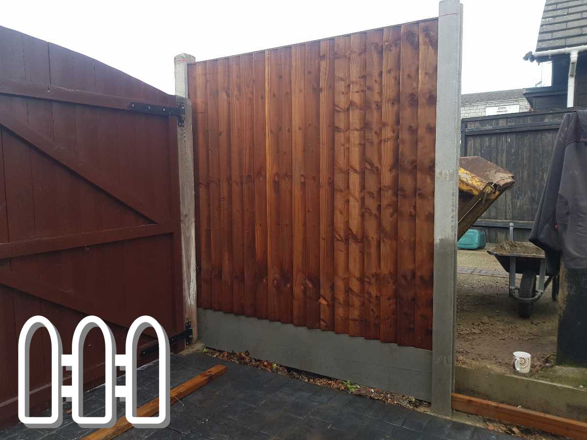 Newly installed wooden fence panel beside a metal gate in an outdoor residential area, with construction tools and a coffee mug visible on the ground.