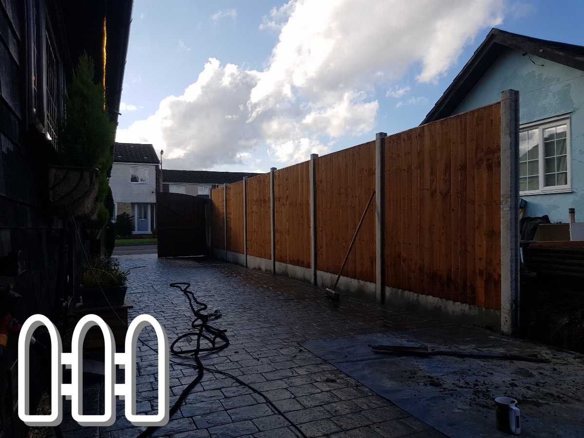 Newly installed wooden fence in a residential driveway, with a view of a house and a paved path, cloudy sky in the background