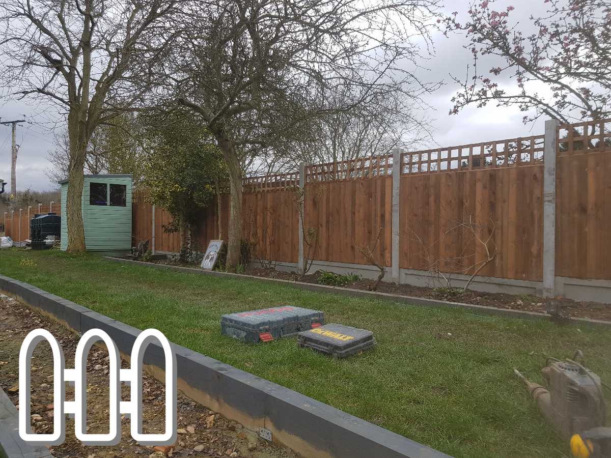 Newly installed wooden fence in a residential backyard with concrete posts and decorative lattice top, accompanied by a small green shed, tools on the grass, and blooming trees.