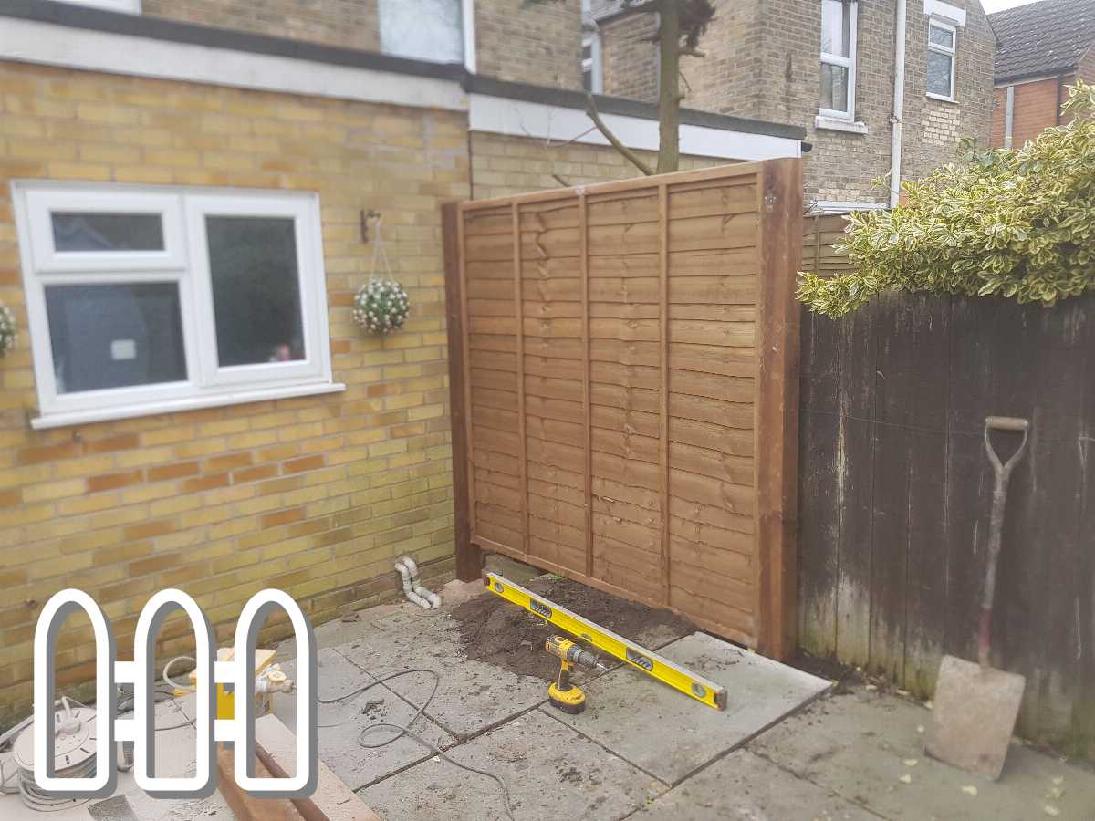 New wooden garden fence installation in progress beside a yellow brick house, featuring tools like a spirit level and electrical saw on a paved area.