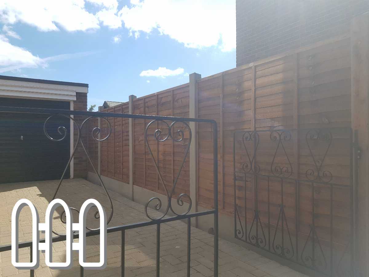 Elegant black wrought iron gate with intricate designs, leading to a residential area with modern wooden fencing and a paved driveway, under a clear blue sky