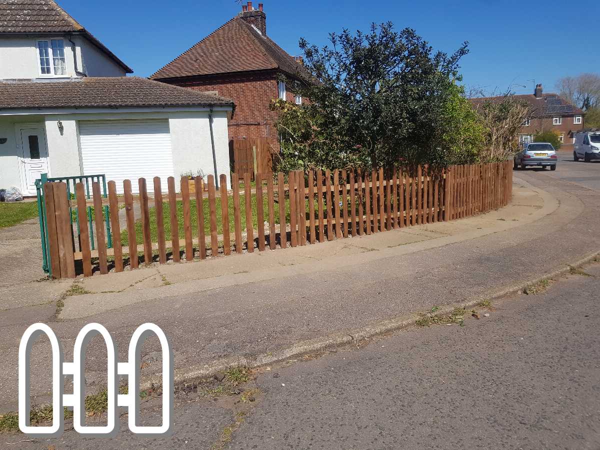 Wooden picket fence in front of a residential property, curving along the sidewalk with a lush green bush behind it, under a clear blue sky