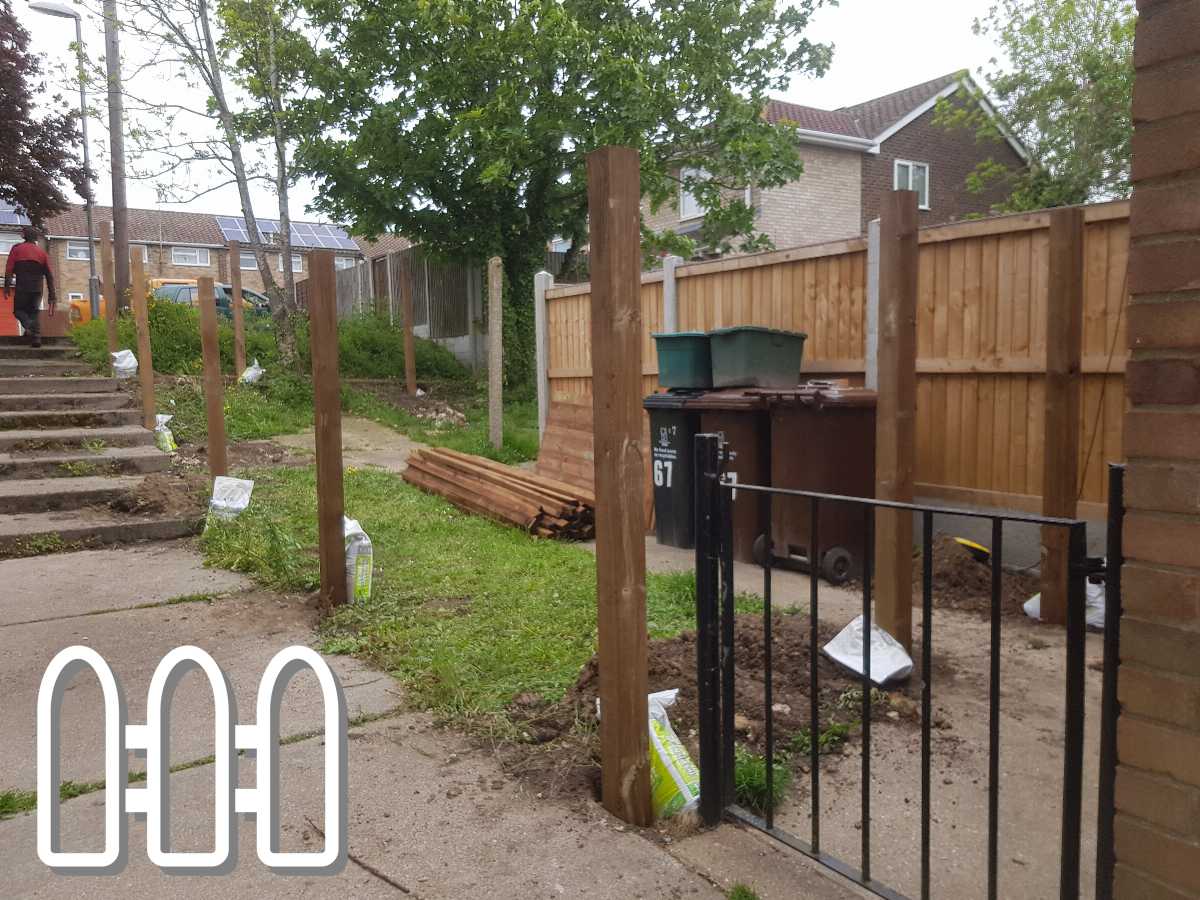 Outdoor construction site showing the early stages of a wooden fence installation with vertical wooden posts in alignment, bags of construction materials on the ground, and a man working in the background.