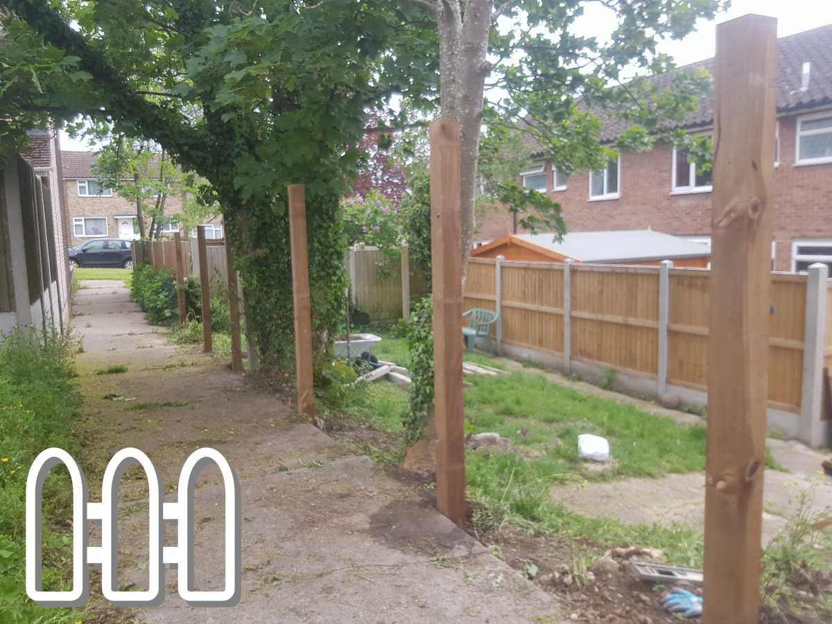 Newly installed wooden fence posts in a residential alleyway with a pathway, overgrown grass, and a view of houses in the background