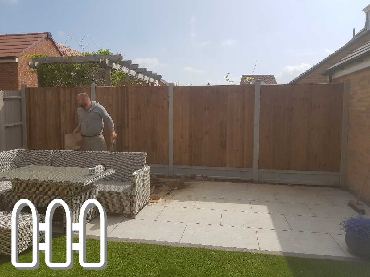 Man installing a wooden fence panel in a sunny, tidy backyard with modern outdoor furniture and finished paving near a brick house.