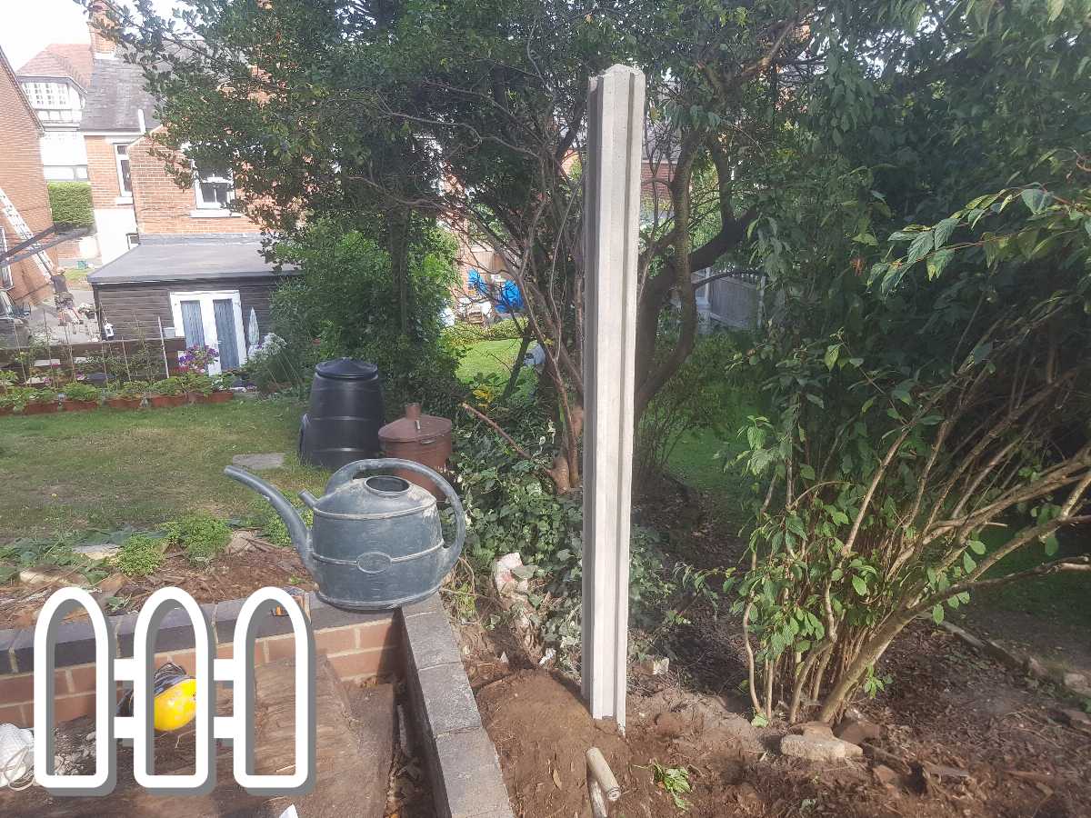 Garden corner with ongoing fence installation, featuring a wooden plank, soil, tools like a yellow helmet and a watering can, surrounded by lush greenery and a distant shed