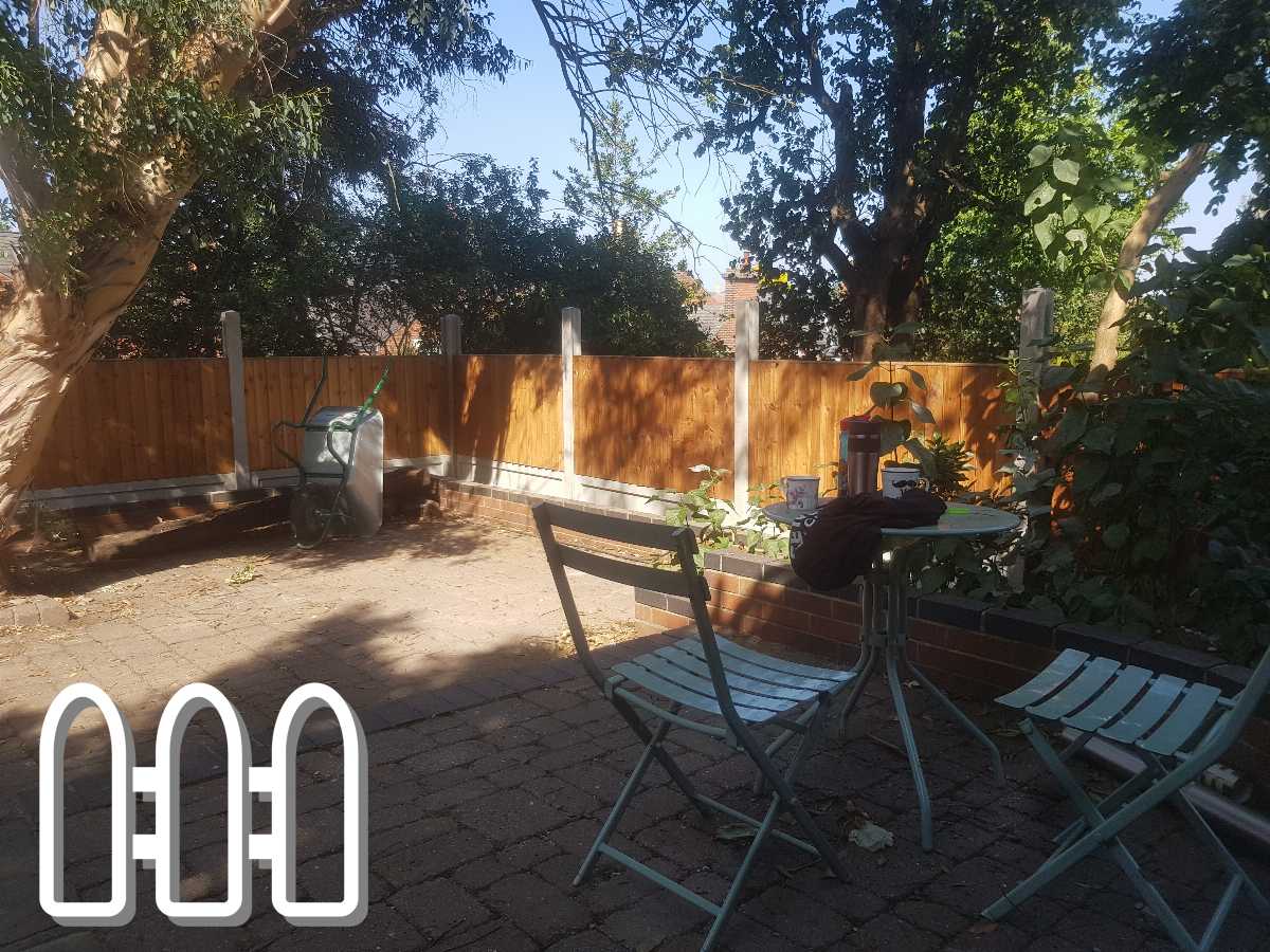 A peaceful backyard garden with a newly installed wooden fence, paving stones, a wheelbarrow, and outdoor dining set under a tree