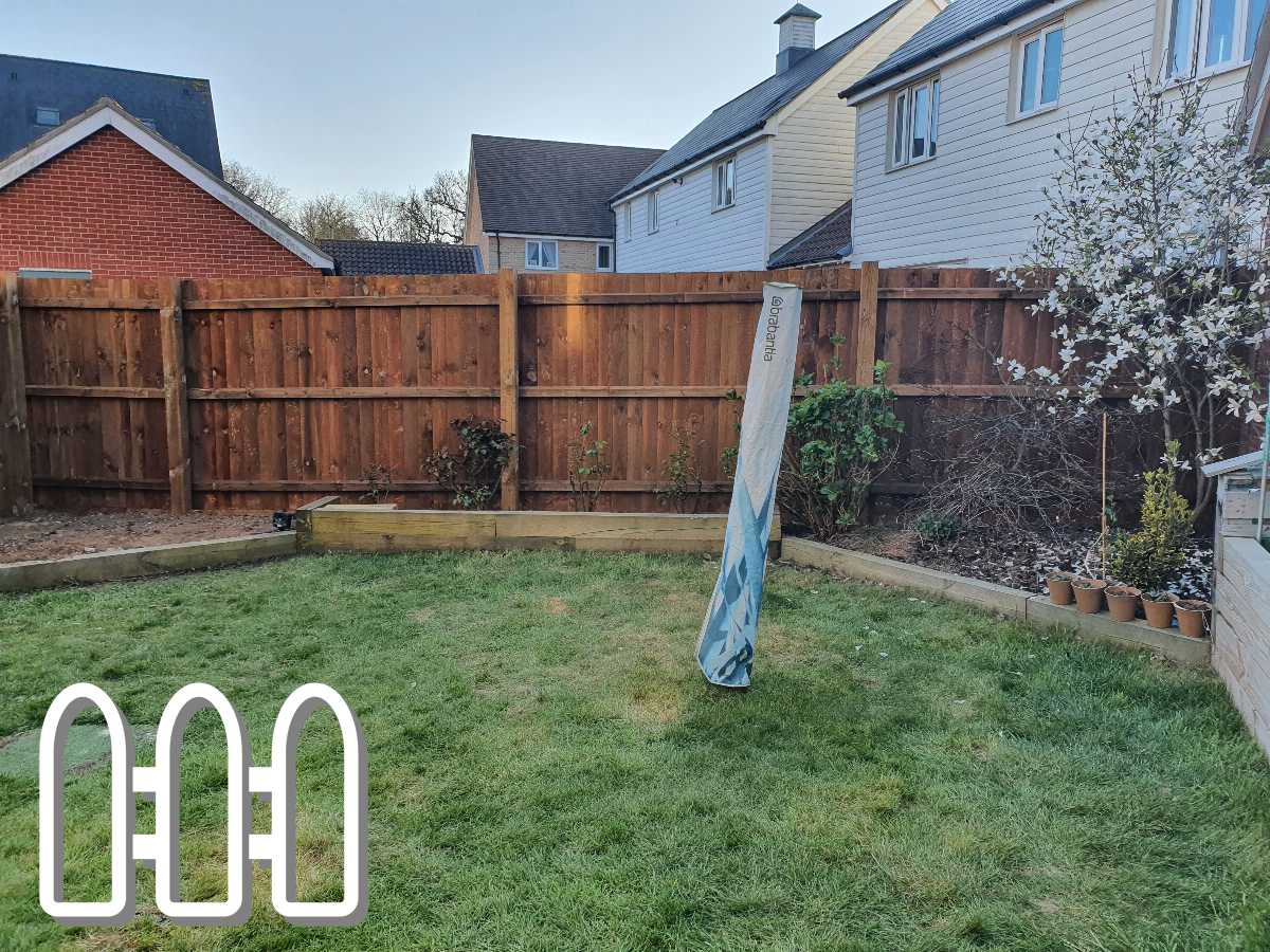 View of a residential garden with a wooden panel fence surrounding the property, featuring a blooming tree and a vertical advertising banner on the grass.