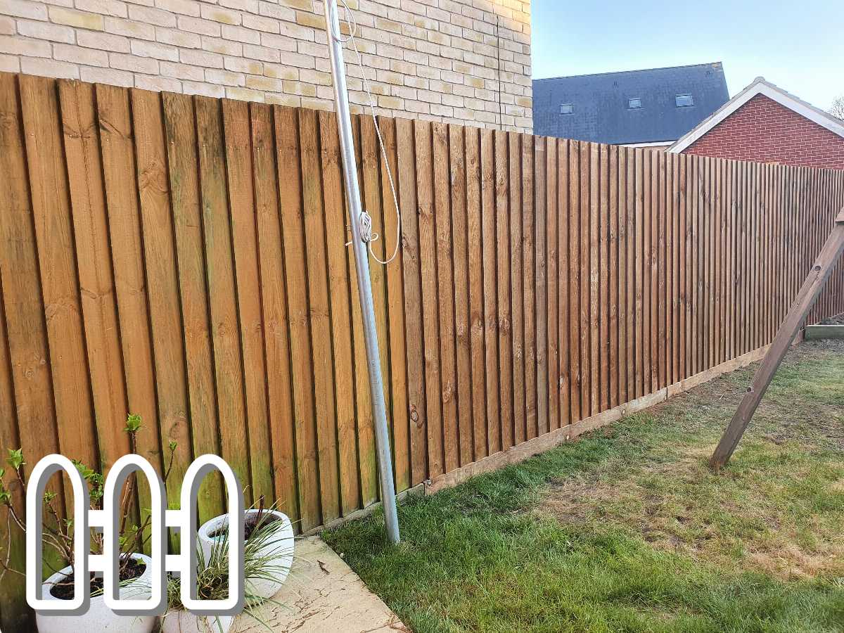 Sturdy wooden fence installation in a residential backyard with additional support beams and lush green grass, featuring plant pots at the base and a partial view of adjoining brick and red-roofed houses.