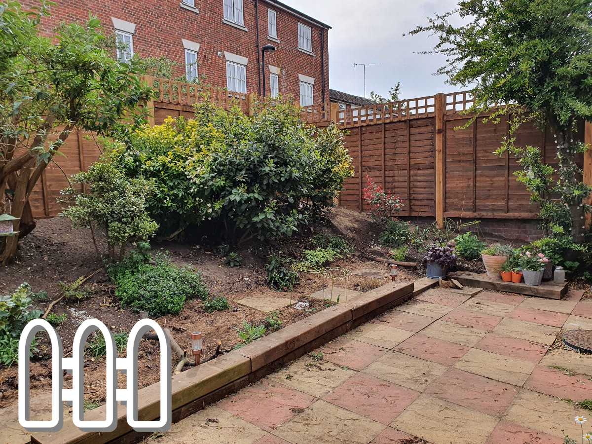 Residential garden under construction with a new wooden fence, planting beds, and various potted plants on a brick patio