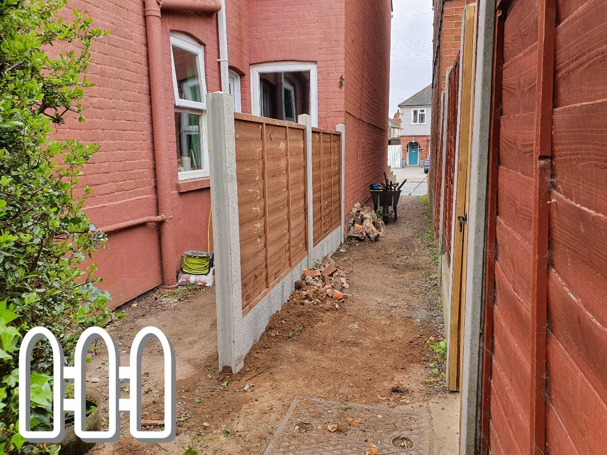 Narrow pathway between a red brick house and a wooden fence, showing recent landscaping work and a wheelbarrow with debris